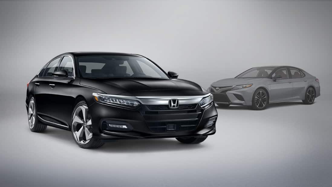 Vista frontal desde el lado del pasajero del Honda Accord Touring 2.0T 2020 estacionado junto a un vehículo Toyota Camry de la competencia.